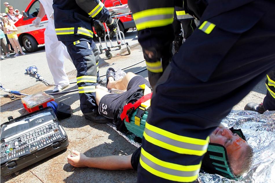 A man is lying on the ground, secured to a backboard, while firefighters in reflective gear provide assistance. Nearby, medical equipment is visible, and an ambulance is parked in the background. Bystanders stand at a safe distance watching the scene unfold, possibly connected to an uninsured motorist claim.